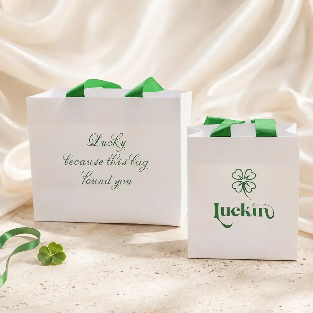 Two Luckin Clover gift bags in ivory with green ribbon, printed with the clover logo and the line 'Lucky because this bag found you'.